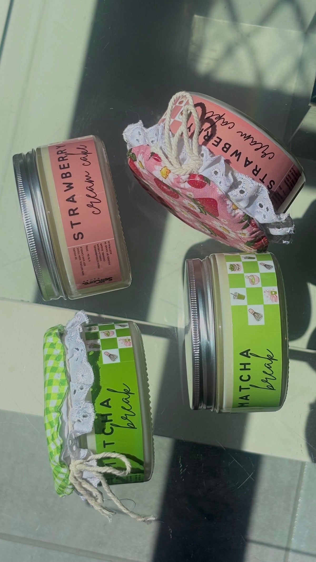 Four small cosmetic jars with metal lids arranged on a glossy glass surface. Two jars have pink labels reading STRAWBERRY, one has a green-and-white check label reading MATCHA, and one has a green-and-white gingham ribbon with lace trim and a white string.