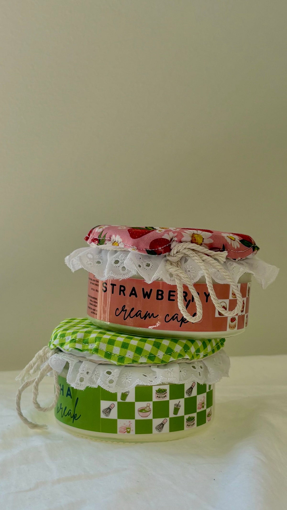 A small decorative jar or container with a pink strawberry-themed lid and lace trim, stacked on top of another container with a green gingham lid, set on a white fabric surface against a plain beige wall.