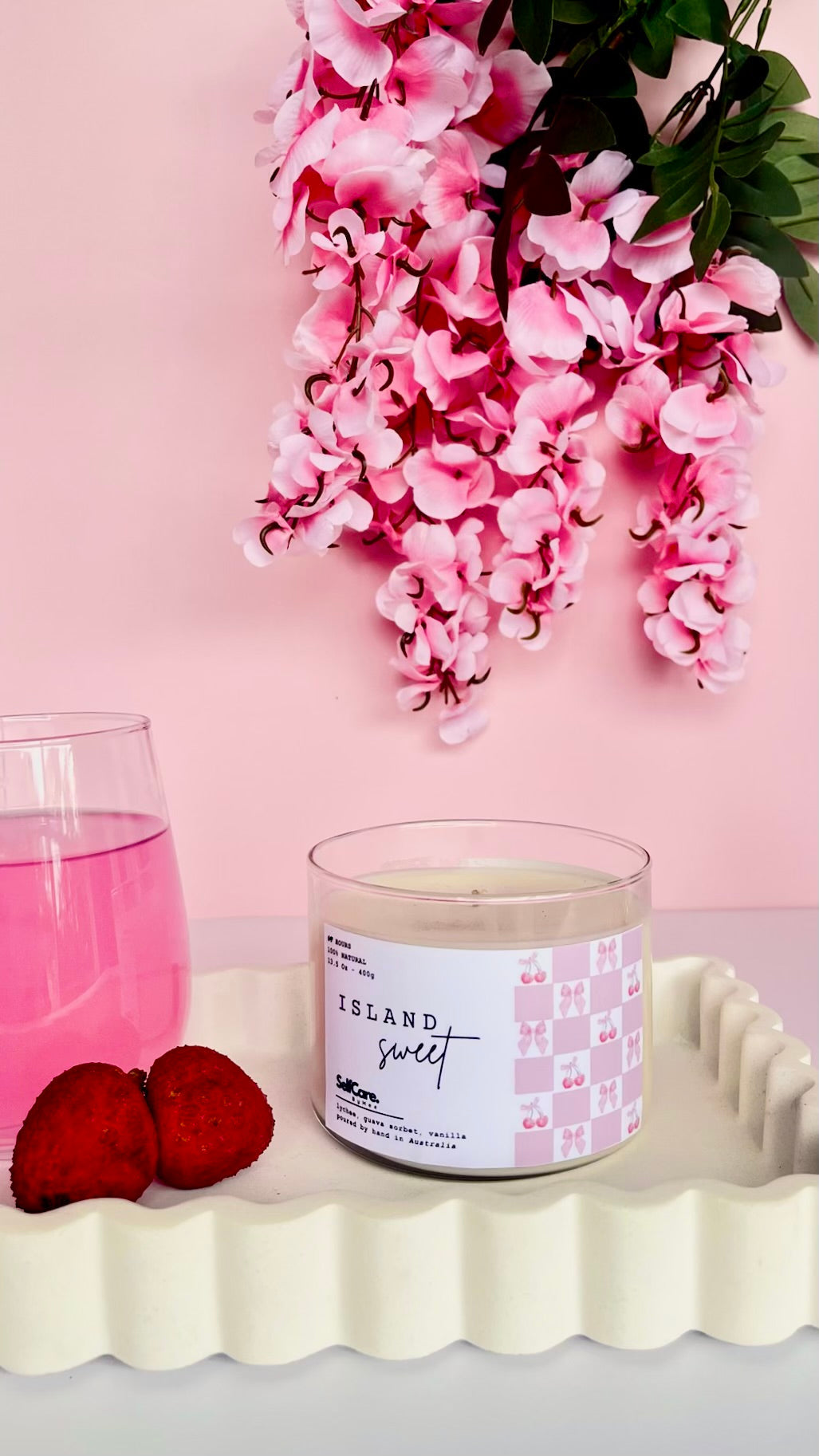 Candle labeled 'Island Sweet' with pink flowers and a glass of pink liquid on a light pink background