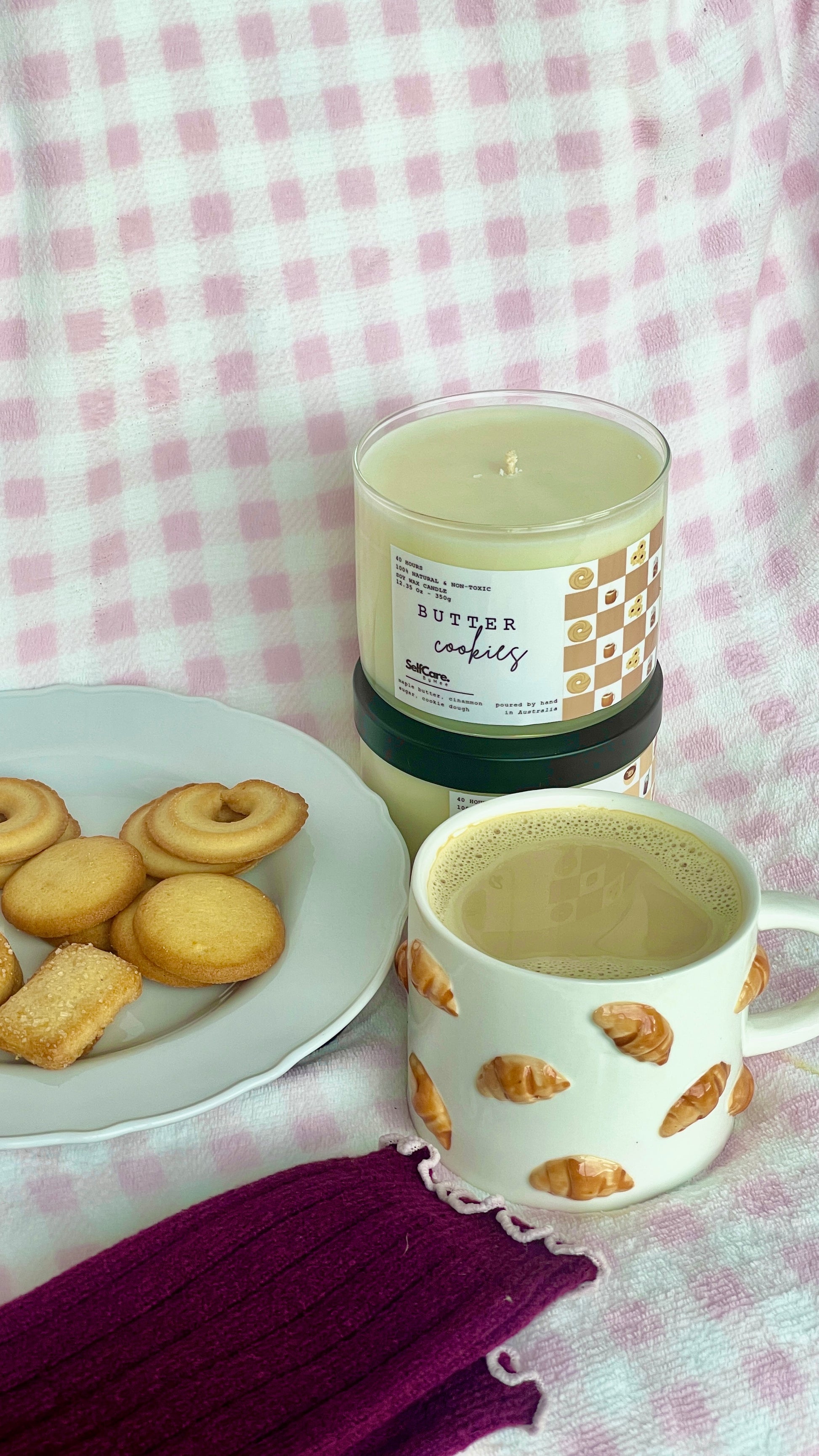 Candle with a honeycomb design next to a mug and cookies on a checkered tablecloth.