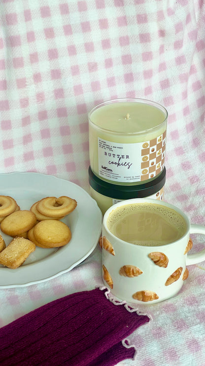 Candle with a honeycomb design next to a mug and cookies on a checkered tablecloth.