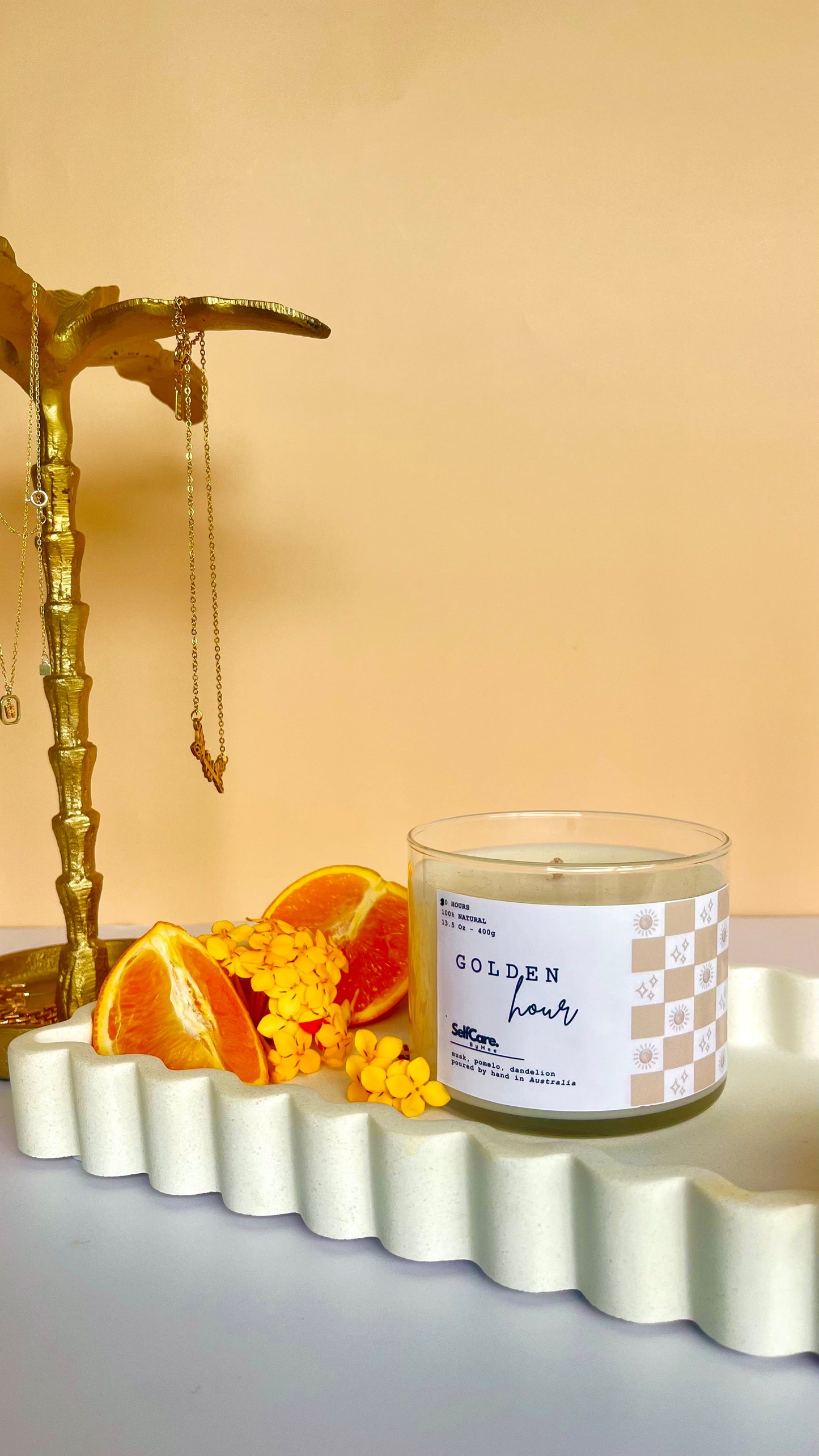 Candle with a label "Golden Hour" on a decorative tray with fruit and flowers against a beige background