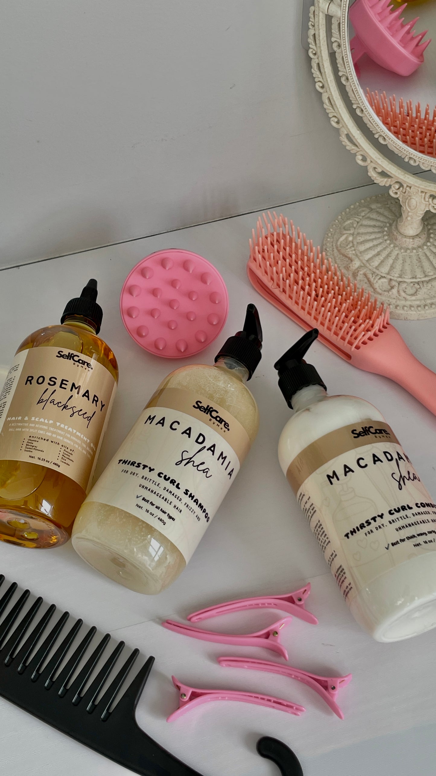 Three bottles of SelfCare by Mee  haircare products (Rosemary Blackseed hair oil, Macadamia Shea Thirsty Curl Cream Shapoo & Conditioner) arranged on a white surface with pink and black hair tools, alongside a mirror reflection.