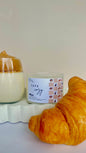 Candle labeled 'Cafe Cozy' with a croissant on a white surface and a coffee latte next to it