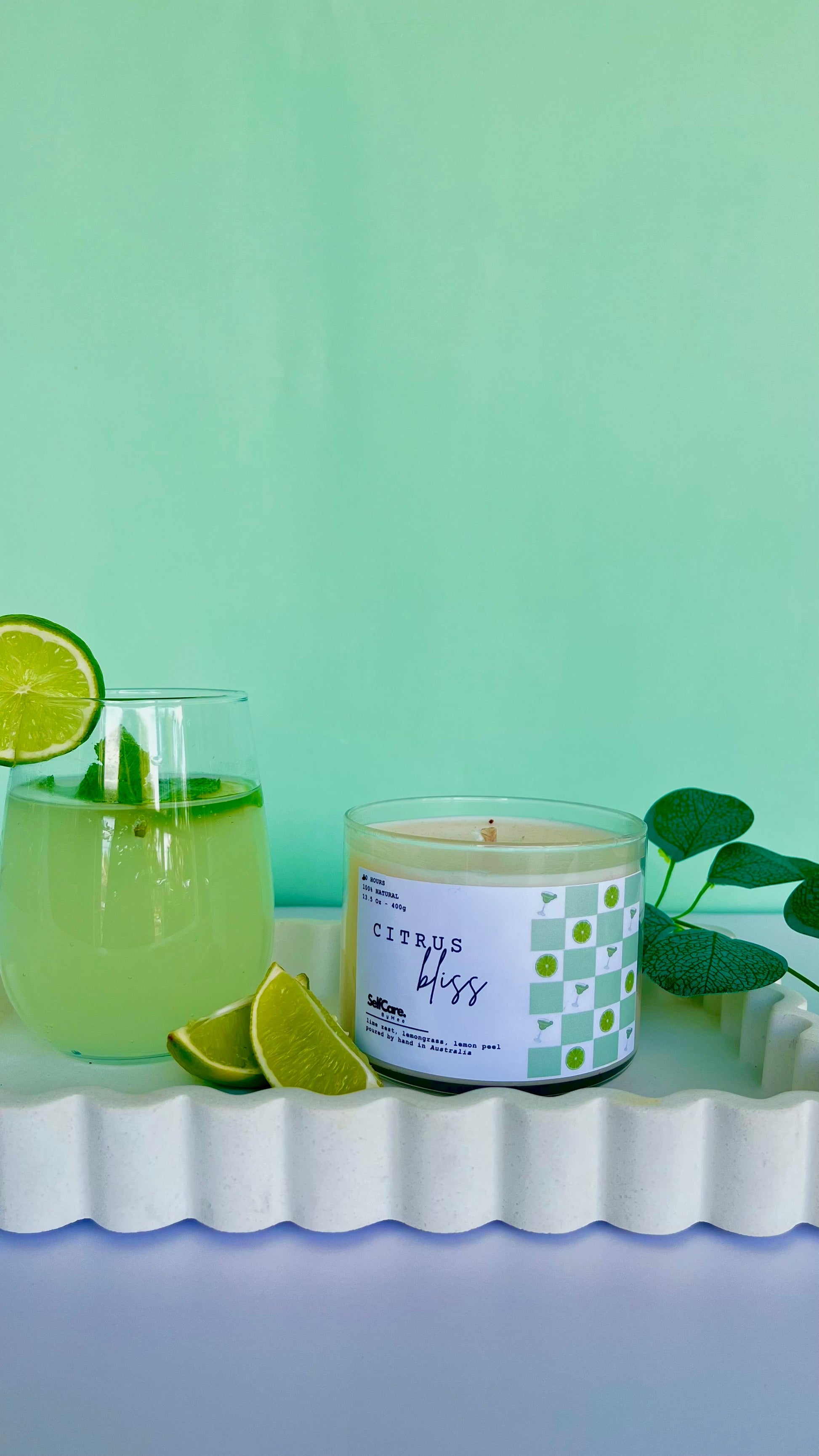 Candle labeled 'Citrus Bliss' next to a glass of lemonade with lime slices on a green background