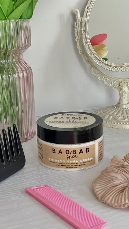 A jar of BAObAB Shea hair moisturizer sits on a light gray tabletop with styled beauty props, including a pink comb, a black hairbrush, a beige scrunchie, and a decorative mirror in the background.