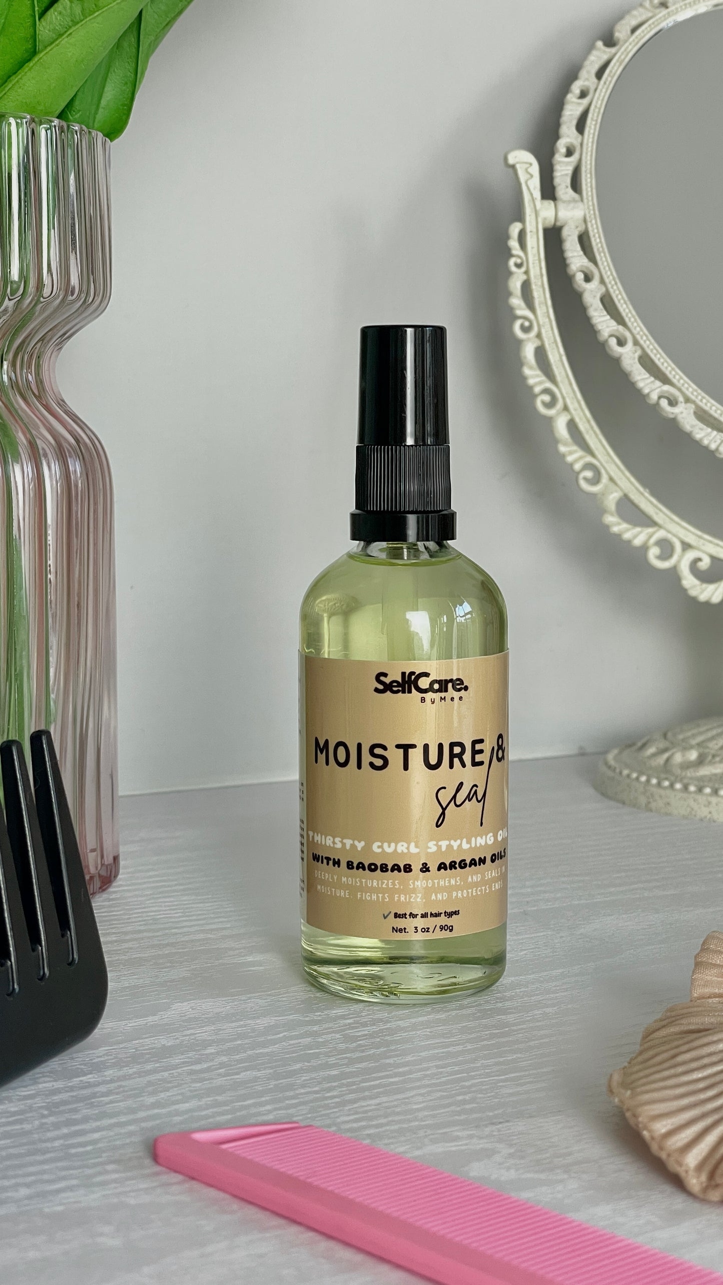 A bottle of SelfCare by Mee Moisture & Seal hair care pump oil with a beige label and black spray cap, set on a light gray textured surface with a pink comb, a pink vase, and a decorative mirror in the background.