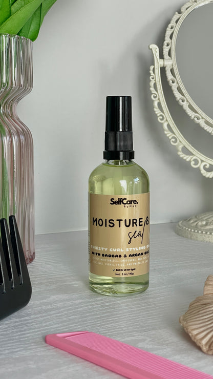 A bottle of SelfCare by Mee Moisture & Seal hair care pump oil with a beige label and black spray cap, set on a light gray textured surface with a pink comb, a pink vase, and a decorative mirror in the background.