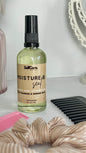 SelfCare by Mee Moisture & Seal thirstly curl styling oil in a pump bottle with a beige label and black pump top, styled on a white surface with a pink comb, black wide tooth hair comb, and beige scrunchie in the foreground