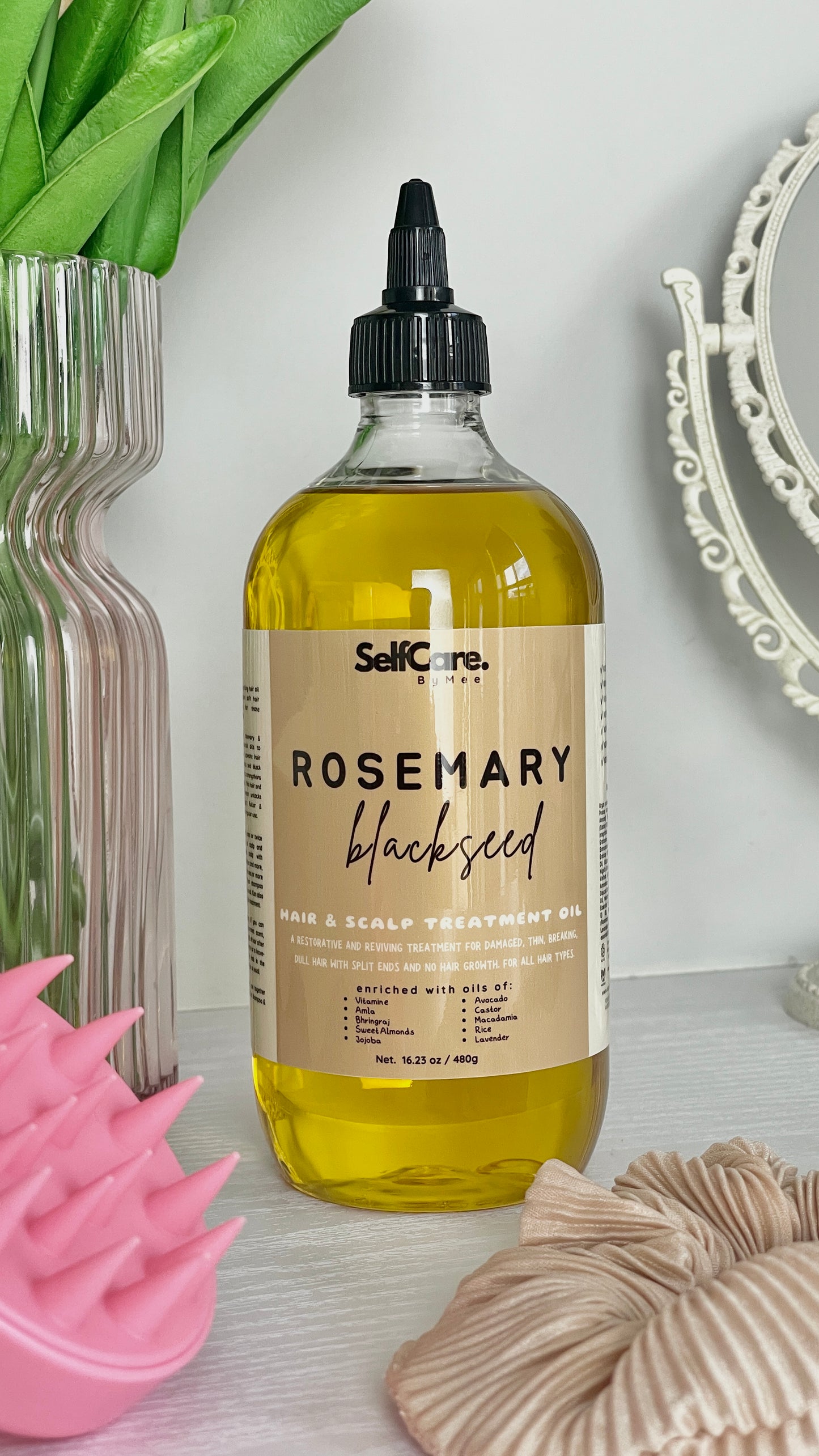 A bottle of SelfCare by Mee Rosemary Blackseed Hair & Scalp Treatment Oil, filled with golden yellow oil, displayed on a light surface with a pink scalp massager, beige scrunchie, and green leaves in the background.
