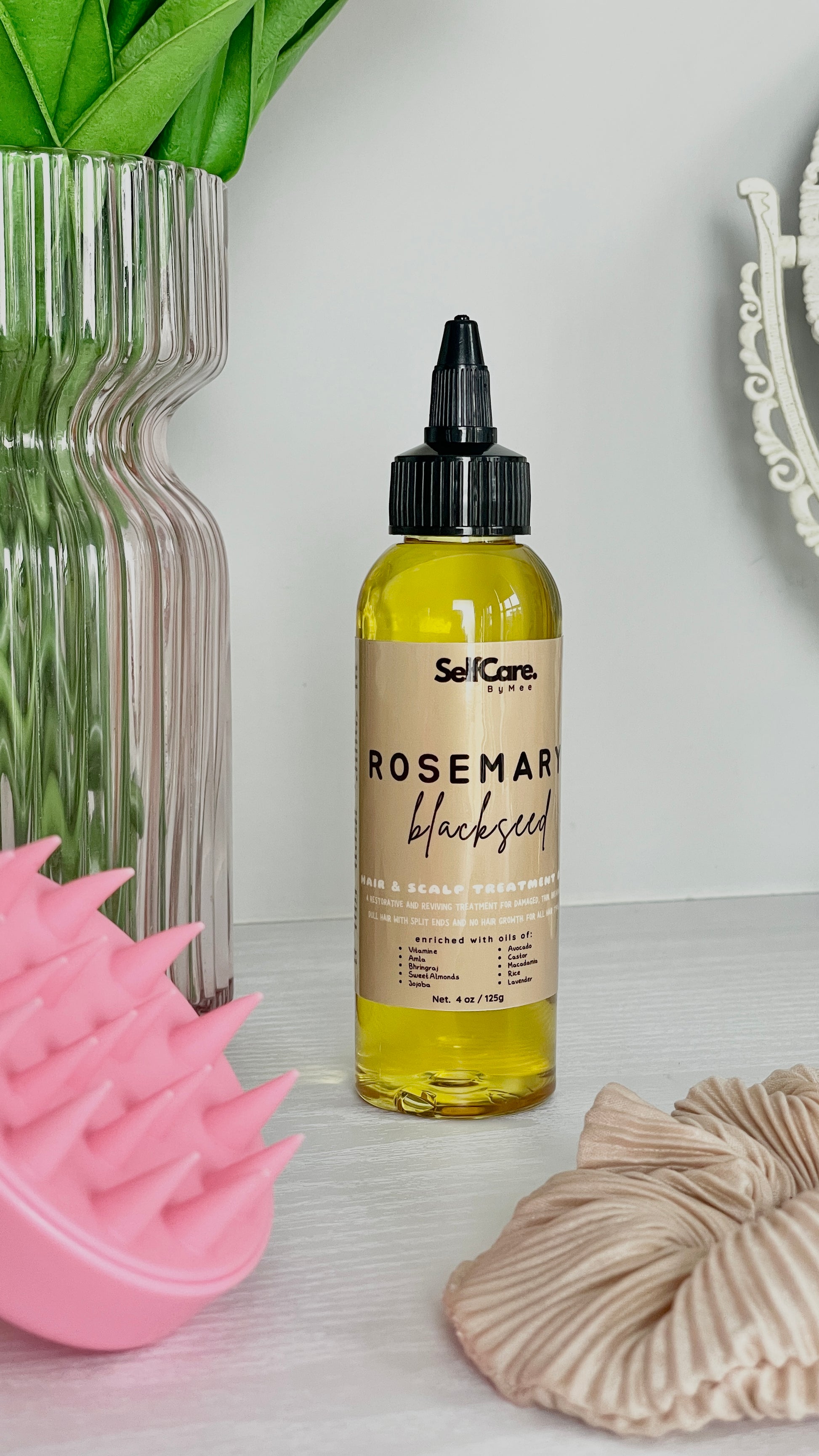 A bottle of SelfCare by Mee Rosemary Blackhead Hair and Scalp Treatment with a black dropper cap, standing on a white surface next to a pink silicone scalp massager, a beige scrunchie, and a glass vase with green leaves.