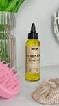 A bottle of SelfCare by Mee Rosemary Blackhead Hair and Scalp Treatment with a black dropper cap, standing on a white surface next to a pink silicone scalp massager, a beige scrunchie, and a glass vase with green leaves.