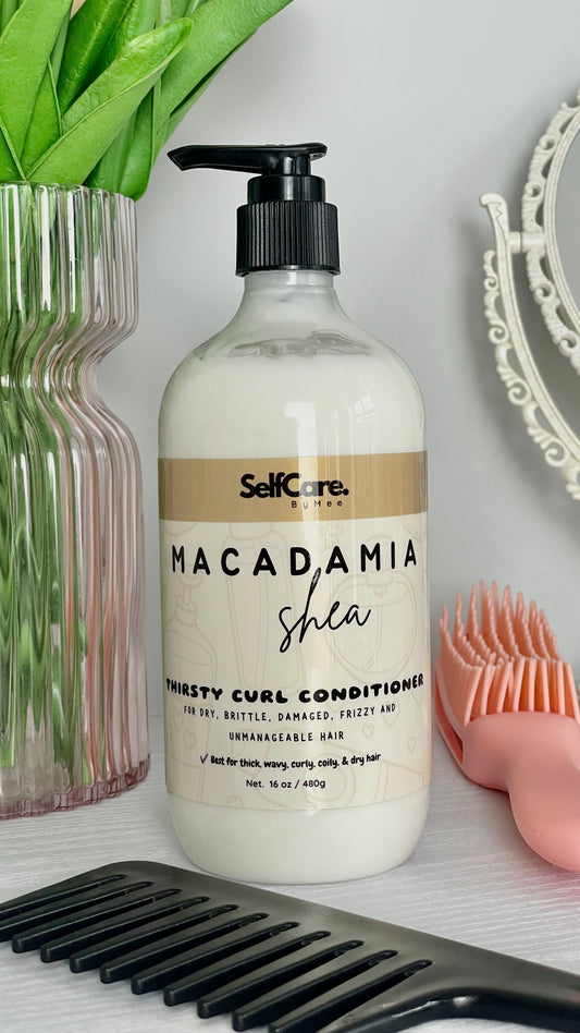 A bottle of SelfCare by Mee Macadamia Shea Thirsty Curl Conditioner stands on a white surface, styled with a black hair comb in front, a pink detangling brush to the right, and a pink ribbed vase with green leaves to the left.