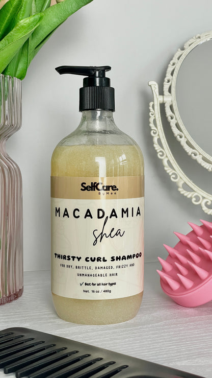 A bottle of SelfCare by Mee Macadamia Shea Thirsty Curl Shampoo stands on a light surface with a pink silicone scalp massager, a decorative mirror, and green leaves in the background.