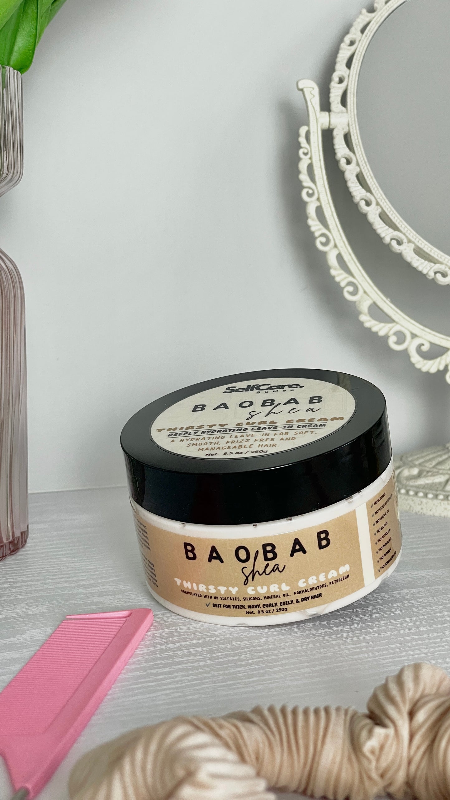 A jar of SelfCare by Mee Baobab Shea Thirsty Curl Cream sits on a light gray surface with a pink comb, beige scrunchie, and a white decorative mirror in the background