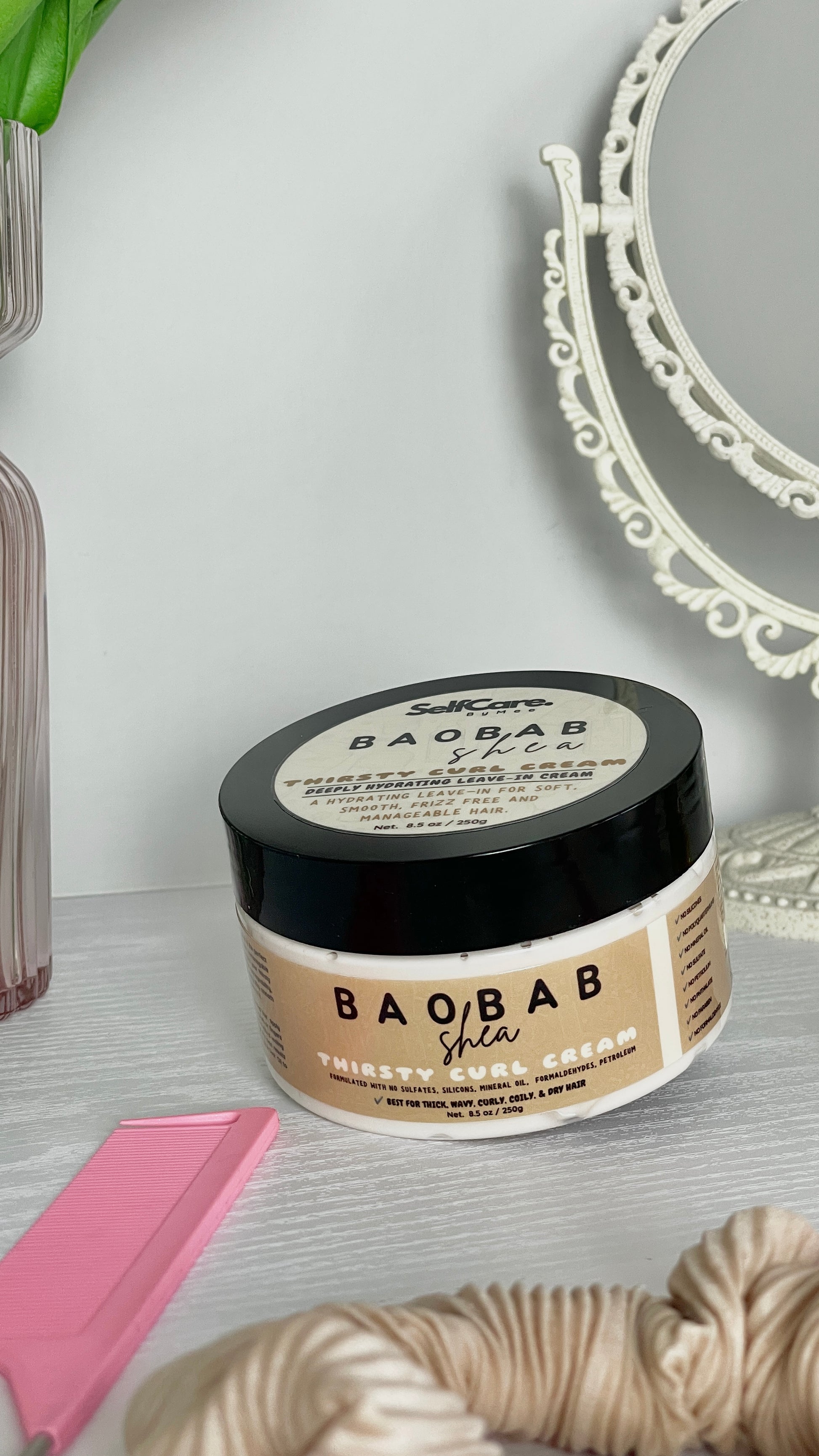 A jar of SelfCare by Mee Baobab Shea Thirsty Curl Cream sits on a light gray surface with a pink comb, beige scrunchie, and a white decorative mirror in the background