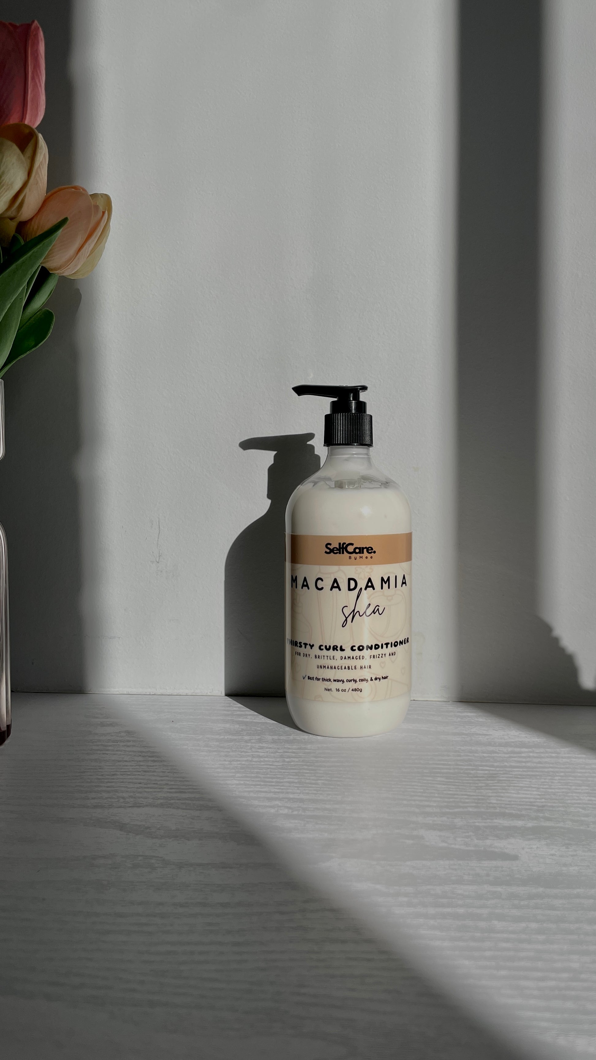 A bottle of SelfCare by Mee Macadamia Shea hair conditioner sits on a light gray wooden surface against a gray wall, with dramatic diagonal shadows and a partial flower arrangement visible on the left edge.