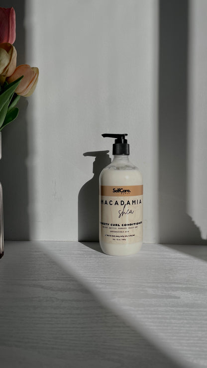A bottle of SelfCare by Mee Macadamia Shea hair conditioner sits on a light gray wooden surface against a gray wall, with dramatic diagonal shadows and a partial flower arrangement visible on the left edge.