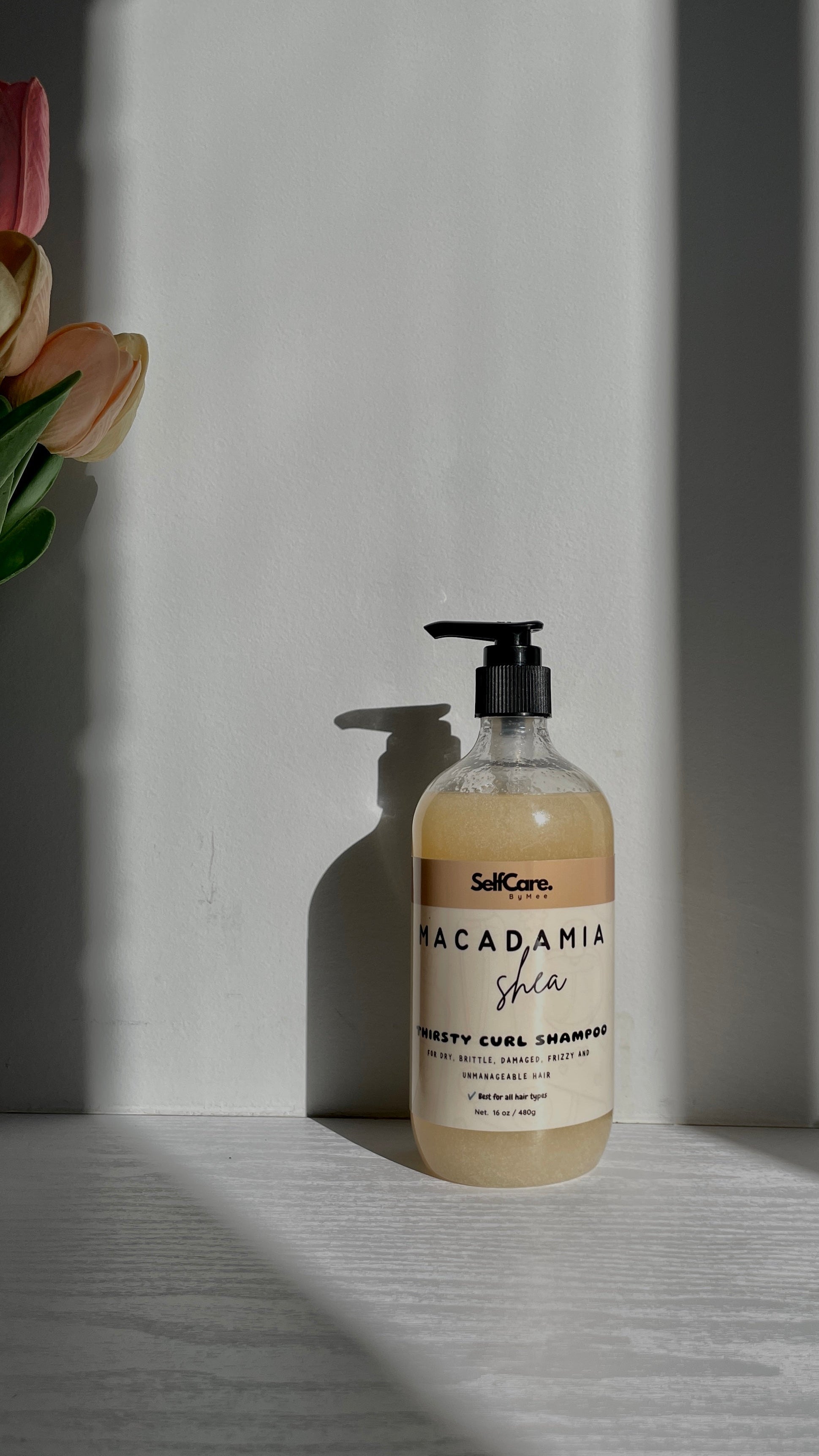 A bottle of SelfCare by Mee Macadamia Shea Hair Cleansing Shampoo with a black pump dispenser, set on a light gray surface with dramatic diagonal shadows and a blurred pink flower in the background.