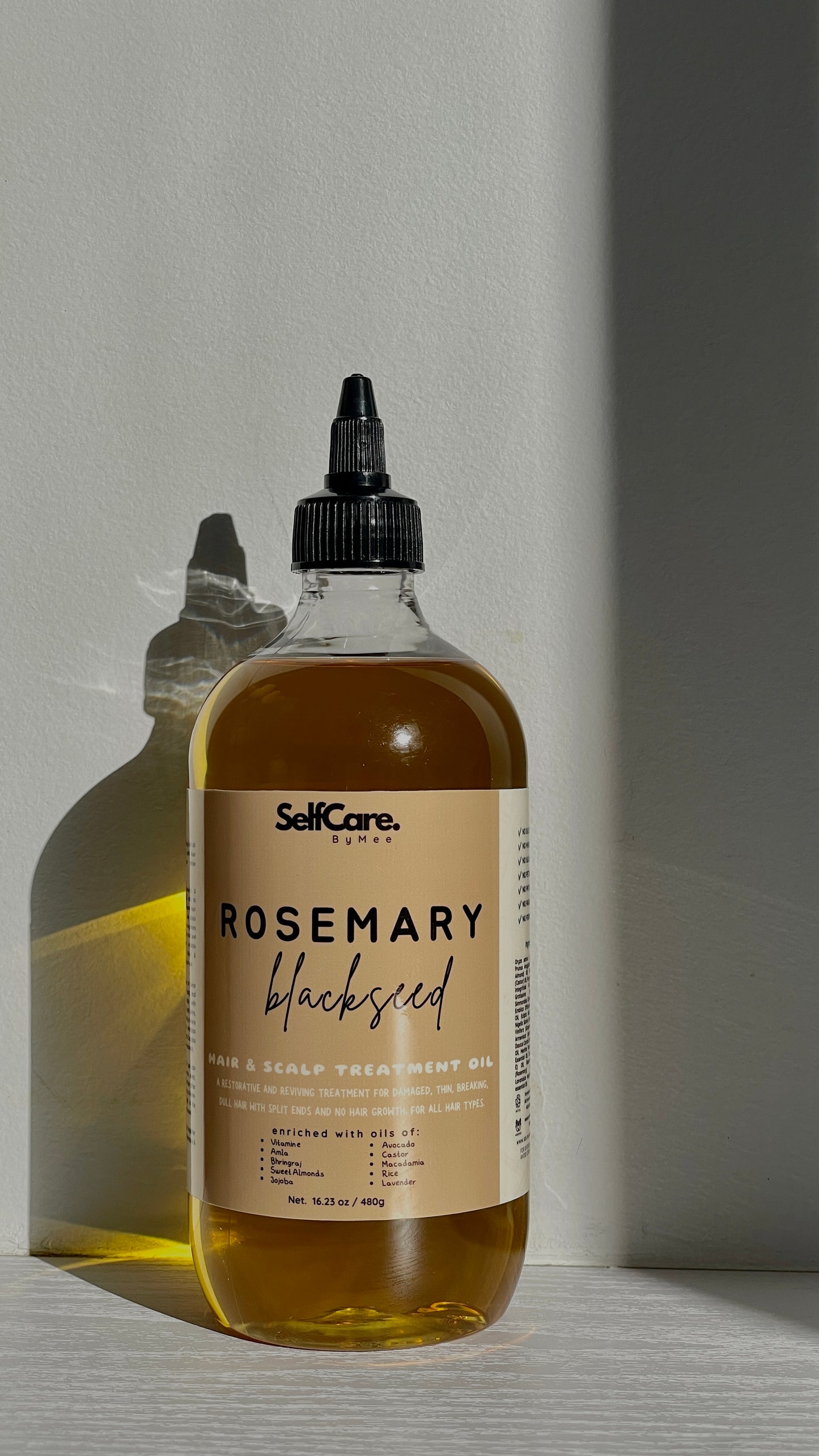 A bottle of SelfCare by Mee Rosemary Blackseed Hair and Scalp Treatment Oil with a black dropper cap, set against a minimalist gray background with dramatic side lighting and a soft reflection.