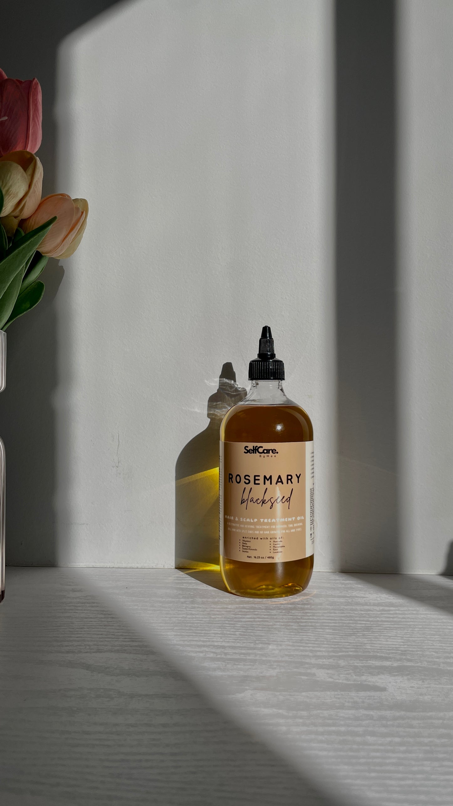 A bottle of SelfCare bt Mee Rosemary Blackseed Hair Oil with a black dropper cap, set on a light gray surface in direct sunlight with strong shadows and a vase of tulips on the left.