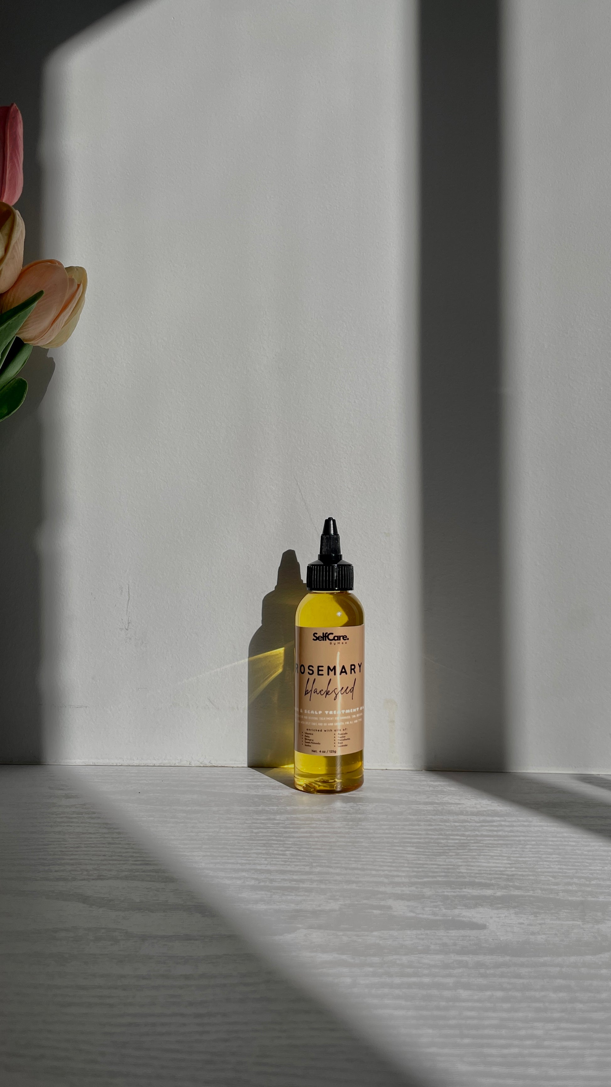 A small glass dropper bottle of rosemary hair oil with a black cap, containing golden-yellow liquid, standing on a light gray surface with dramatic diagonal shadows cast across a white wall and tabletop.