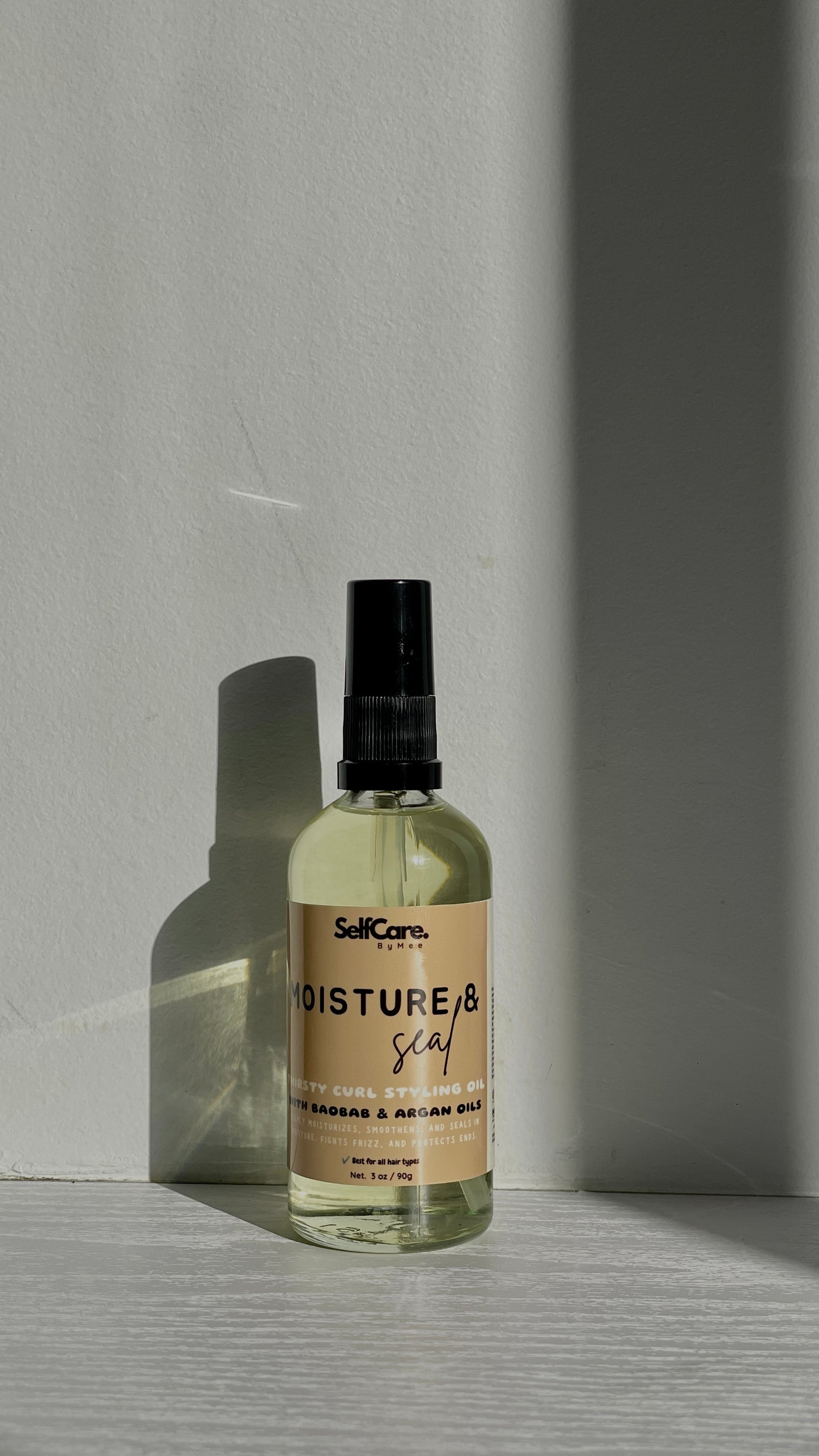 A clear spray bottle with a black nozzle and a beige label reading 'SelfCare by Mee Moisture & seal' stands on a light surface against a gray wall, lit by directional sunlight that casts a strong shadow.