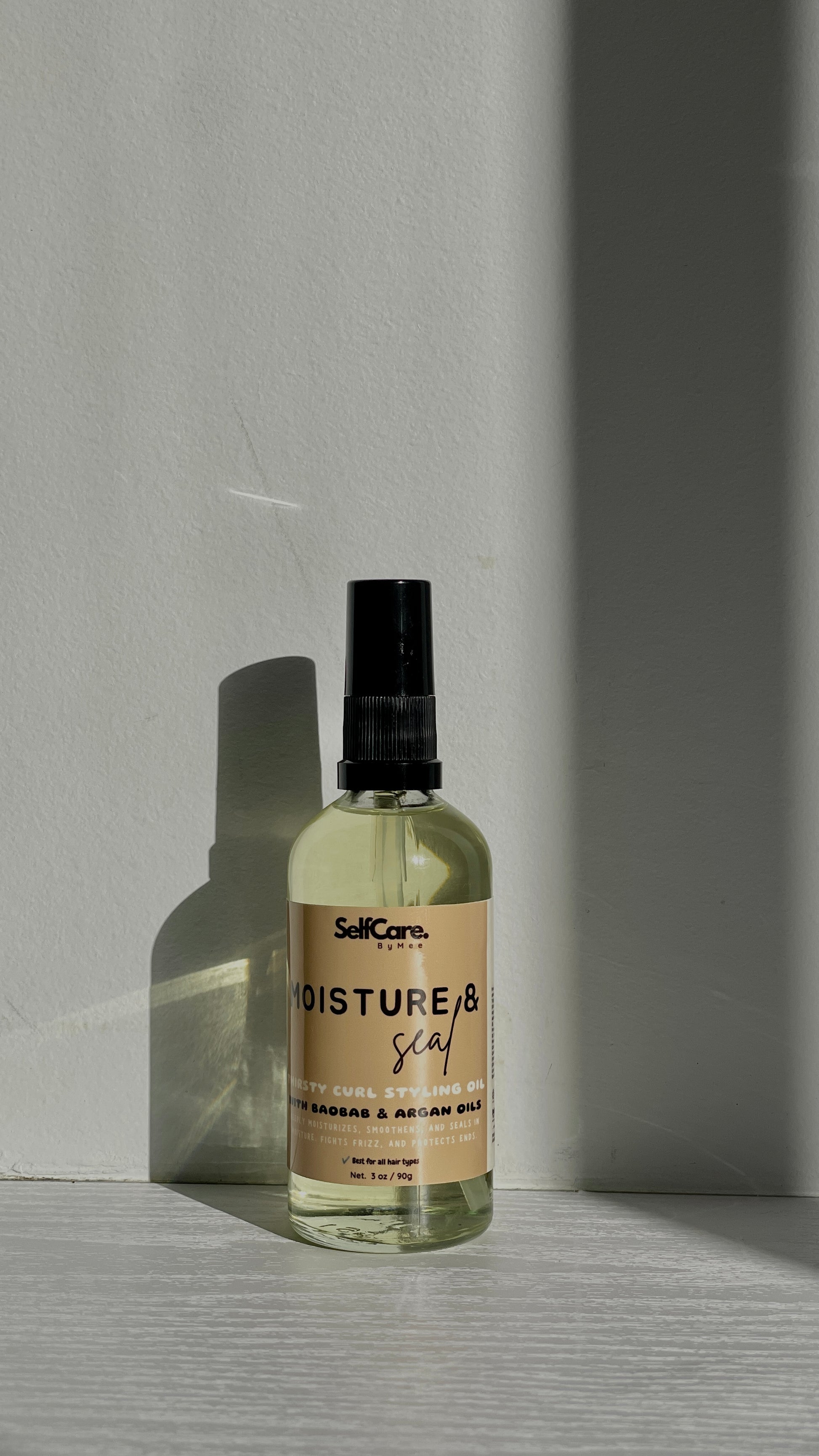 A clear spray bottle with a black nozzle and a beige label reading 'SelfCare by Mee Moisture & seal' stands on a light surface against a gray wall, lit by directional sunlight that casts a strong shadow.