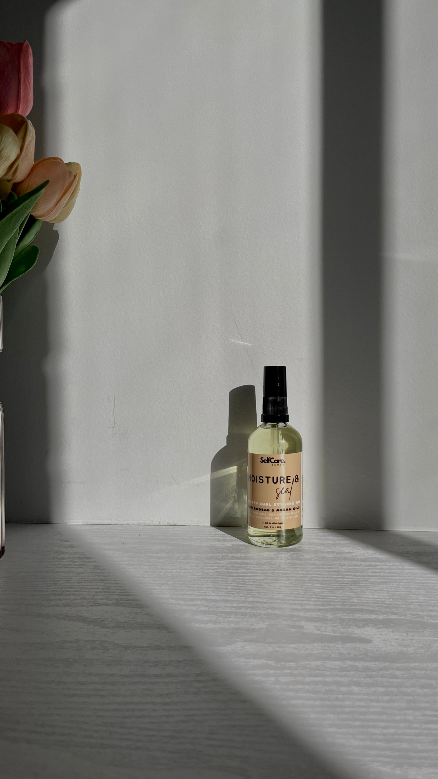 A small clear glass bottle with a black spray top and a cream label reading 'SelfCare by Mee Moistur & Seal thirsty curl styling oil'  sits on a light gray surface in sharp sunlight with strong shadows. A vase with pink and yellow tulips is partially visible on the left.