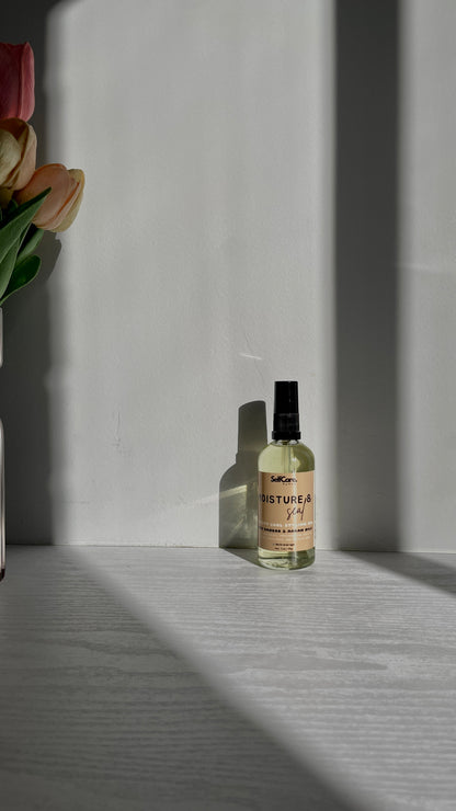 A small clear glass bottle with a black spray top and a cream label reading 'SelfCare by Mee Moistur & Seal thirsty curl styling oil'  sits on a light gray surface in sharp sunlight with strong shadows. A vase with pink and yellow tulips is partially visible on the left.