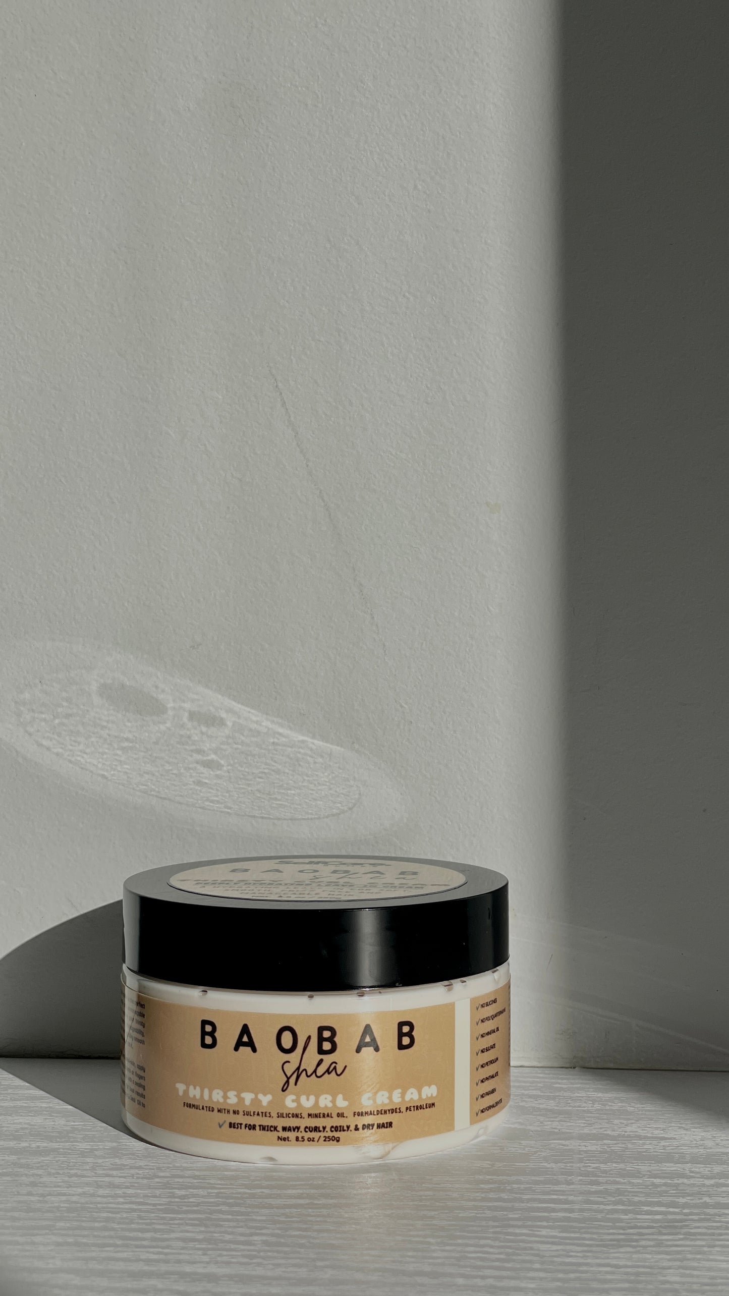 A jar of BAOBAB Shea Thirsty Curl Cream resting on a light gray wooden surface with dramatic diagonal shadows cast across the background wall.