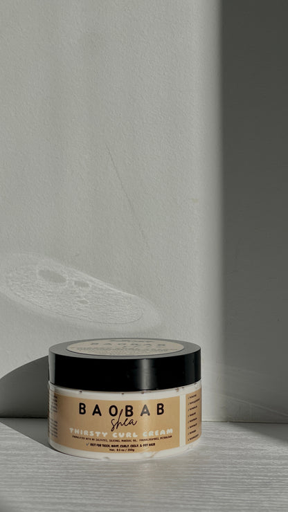 A jar of BAOBAB Shea Thirsty Curl Cream resting on a light gray wooden surface with dramatic diagonal shadows cast across the background wall.