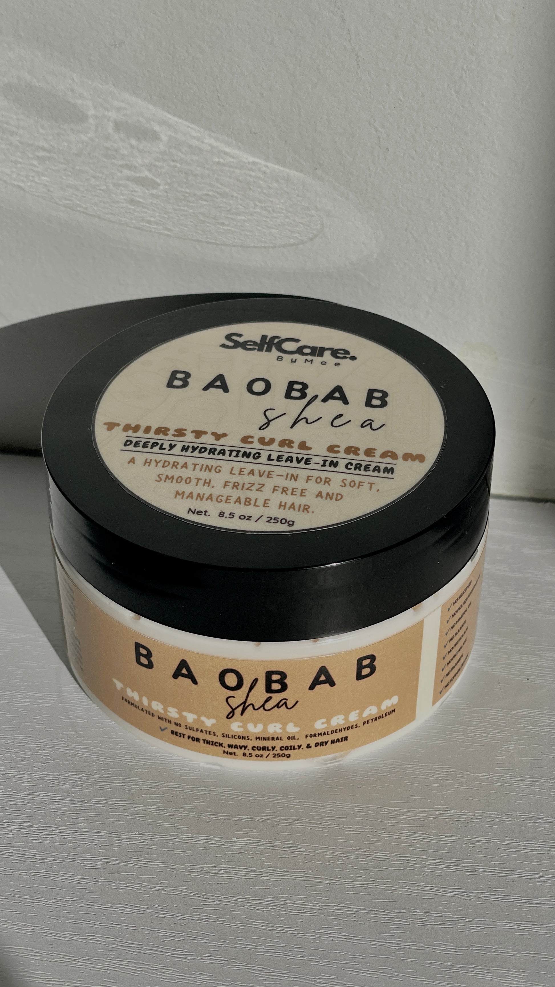 A jar of SelfCare by Mee Baobab Shea Thirsty curl Leave-In Cream with a black lid and beige label, photographed on a light textured surface with a soft shadow.
