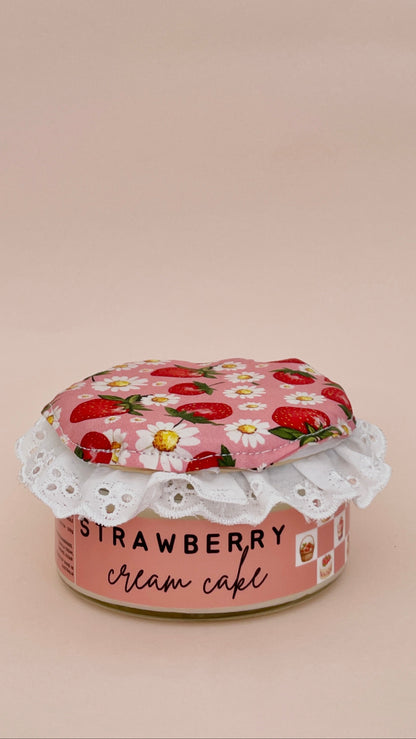 A small pink canister labeled 'STRAWBERRY cream cake' with a strawberry-and-white flower fabric lid and white lace trim, photographed on a plain warm beige background.