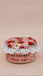 A small pink canister labeled 'STRAWBERRY cream cake' with a strawberry-and-white flower fabric lid and white lace trim, photographed on a plain warm beige background.