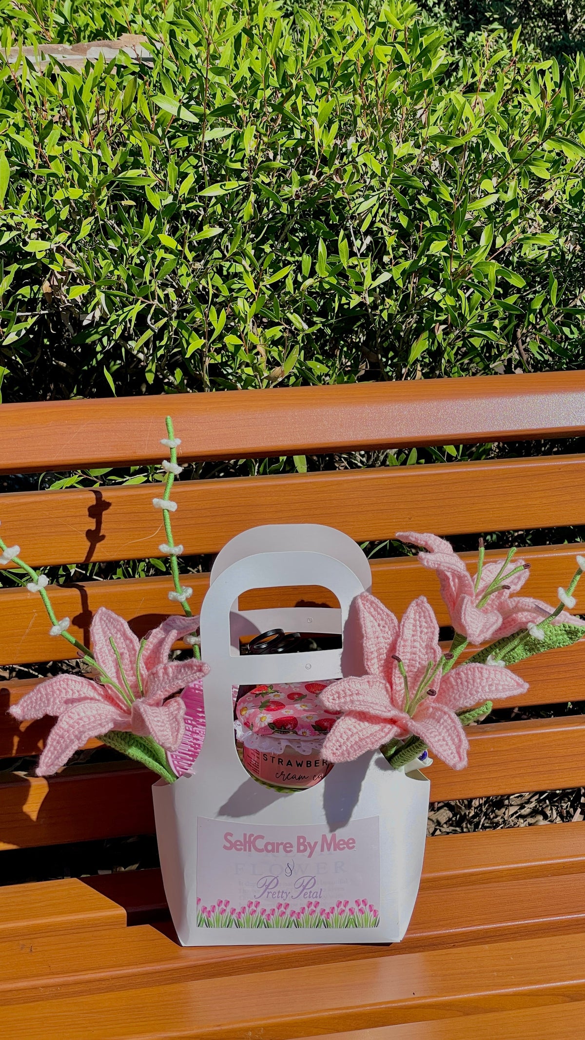 A white paper gift bag with pink floral accents sits on a wooden bench outdoors, with green foliage in the background. The bag has the text 'SelfCare By Mee' printed on the front.