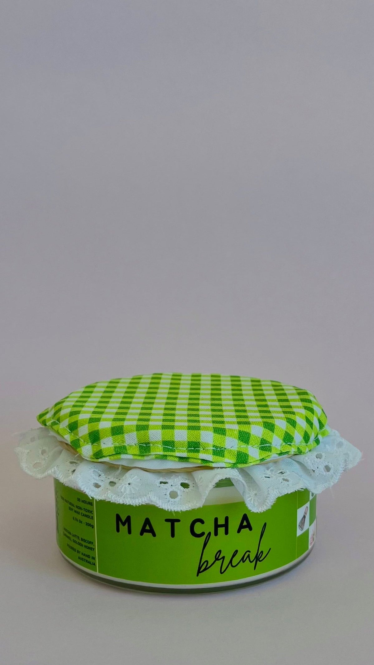 A small jar of matcha coffee with a green and white checkered fabric lid and a white lace trim, set against a plain light gray background.