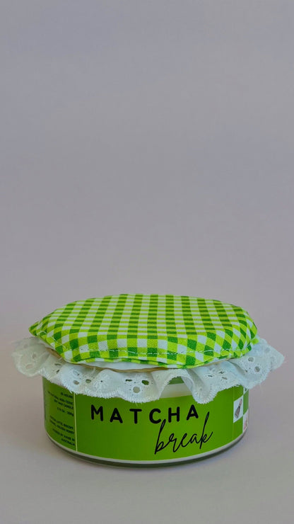 A small jar of matcha coffee with a green and white checkered fabric lid and a white lace trim, set against a plain light gray background.