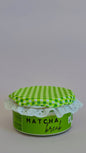 A small jar of matcha coffee with a green and white checkered fabric lid and a white lace trim, set against a plain light gray background.