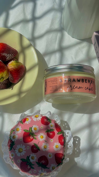 A styled flat-lay featuring a clear jar of 'Strawberry Cream Cake' with a pink label, a pink floral cake topper, and fresh strawberries on a pale yellow plate, all set on a white surface with dappled sunlight and shadows.